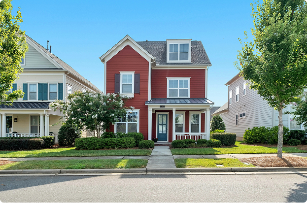 A two-story red house with white trim, a covered front porch, and a small front yard, situated between similar neighboring homes on a sunny day.