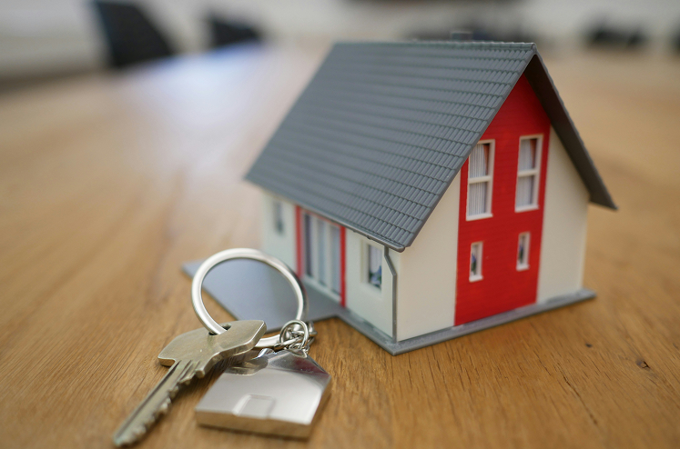 A set of house keys with a house-shaped keychain lies on a wooden table next to a small model house with a red and white exterior.