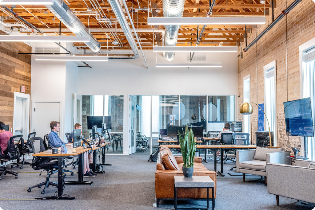 Modern open-plan office with people working at desks, large windows, exposed ceiling pipes, a brown leather sofa, and potted plants.