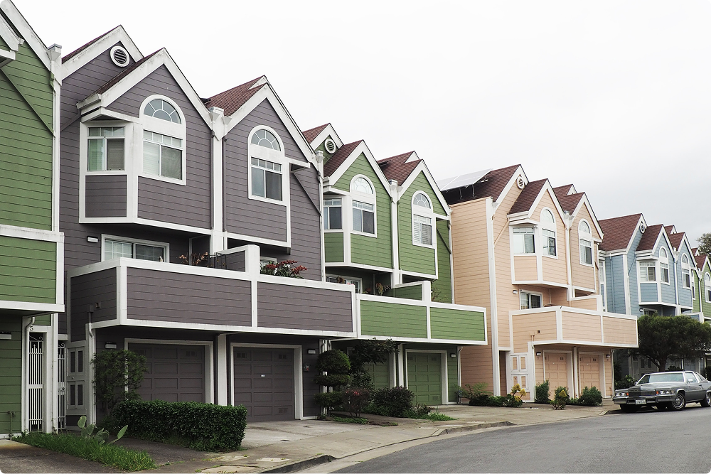 A row of modern three-story townhouses in various colors, featuring garages, balconies, and large windows, lined up along a residential street.