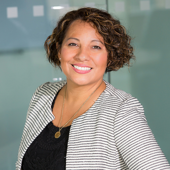 A woman with short curly hair, wearing a striped blazer and black top, stands smiling in front of a glass wall.