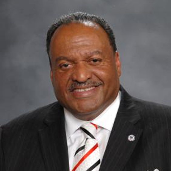 A man in a dark suit, white shirt, and striped tie is smiling at the camera with a gray background behind him.