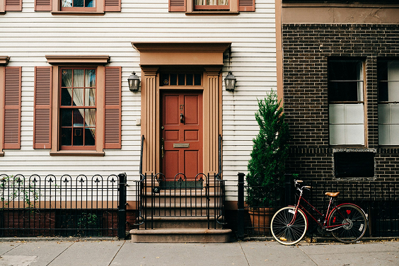 A beige house with brown shutters and door, next to a brown brick wall. A black metal fence and a bicycle are in front of the entrance.