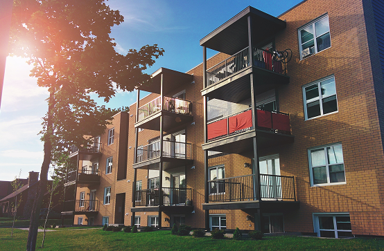 A three-story brick apartment building with balconies and large windows, surrounded by a grassy lawn and trees, under a clear blue sky with sunlight.