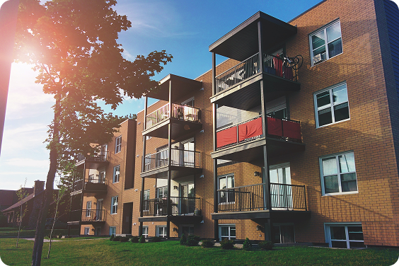 A three-story brick apartment building with balconies and large windows, surrounded by a grassy lawn and trees, under a clear blue sky with sunlight.