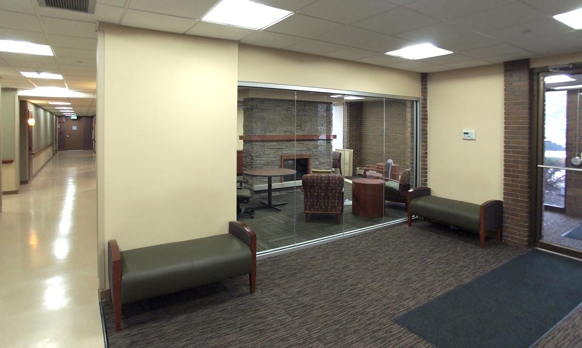 A hallway with two benches leads to a glass-walled lounge area with armchairs, tables, and a brick fireplace visible inside.