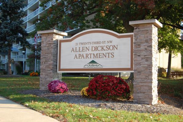 Sign reading "Allen Dickson Apartments" with address, surrounded by flowers and grass, in front of a multi-story building and trees.