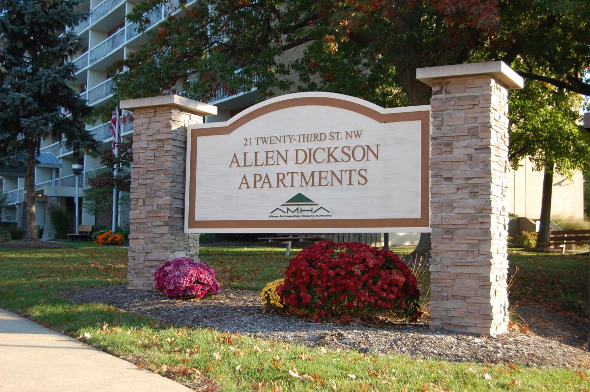 A sign for Allen Dickson Apartments at 21 Twenty-Third St. NW stands in front of a building, surrounded by grass, flowers, and trees.