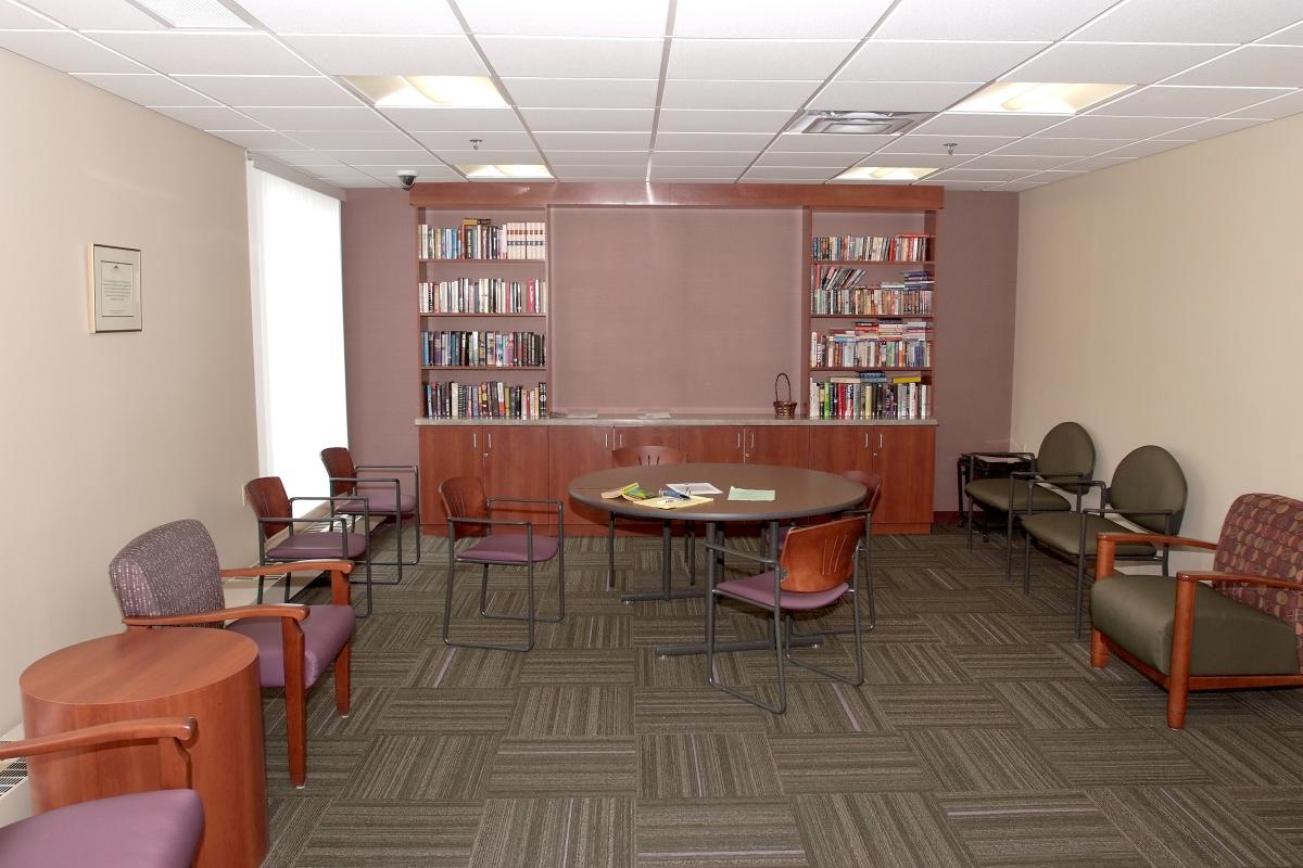 A small meeting room with a round table, several chairs, bookshelves filled with books, and fluorescent ceiling lights.
