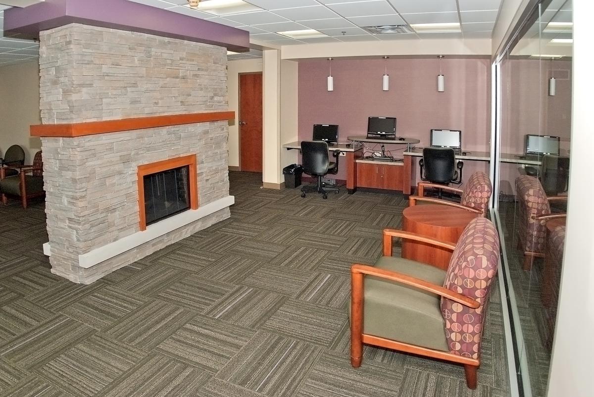 A lounge with a stone fireplace, four desktop computers on a counter, office chairs, and upholstered armchairs on patterned carpet.