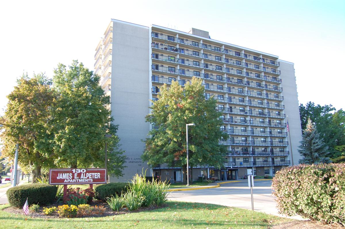 A tall apartment building with balconies, trees in front, and a sign reading “130 James E. Alpeter Apartments” near the entrance.