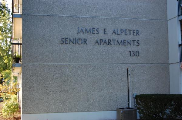 Exterior wall of a building with the sign "James E. Alpeter Senior Apartments 130" in block letters. Bushes and part of an air conditioning unit are visible in front.