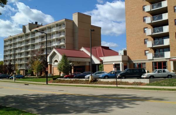 A street view of two mid-rise apartment buildings, a parking lot with several cars, and a clear sky with scattered clouds.