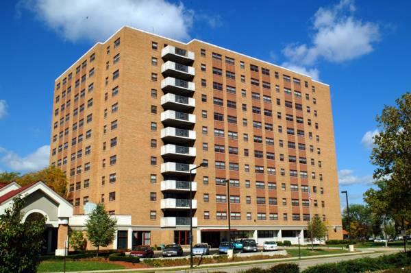 A multi-story brick apartment building with balconies, parked cars in front, and trees nearby under a blue sky with scattered clouds.