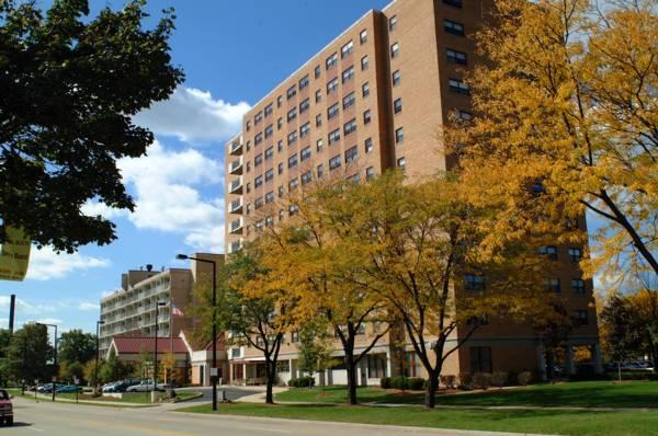 A multi-story brick apartment building stands behind trees with yellow autumn leaves under a partly cloudy sky.