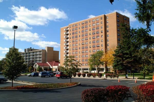 A mid-rise apartment building with balconies stands near a parking lot with cars, surrounded by trees and landscaped bushes under a partly cloudy sky.