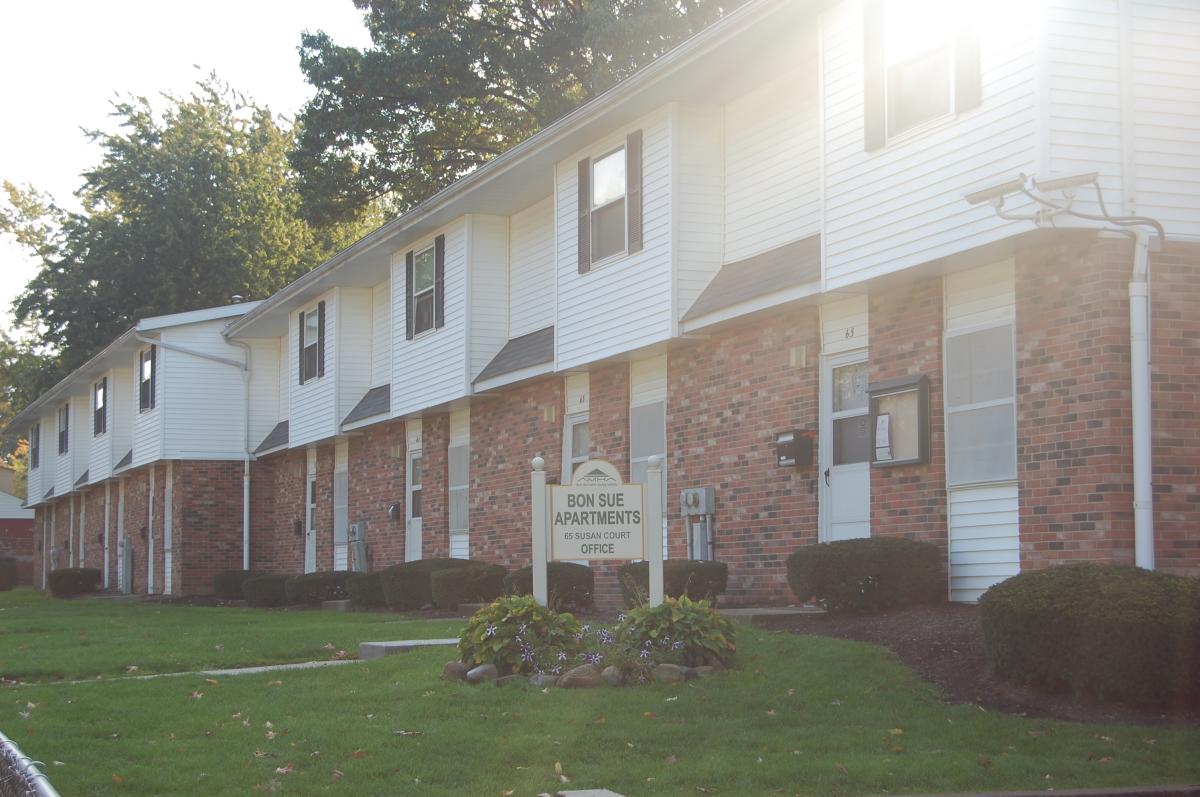 A row of two-story apartment units with brick and white siding, green lawns, and a sign reading "Bon Sue Apartments & Bristol Court Office.