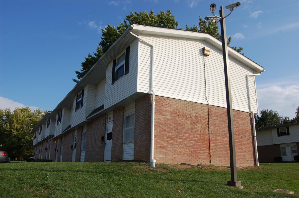 Two-story apartment building with brick lower level, white upper level, and a security camera pole on a grassy lawn under a blue sky.