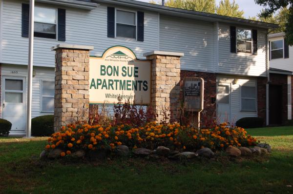 Bon Sue Apartments sign with flower bed in front, situated on a lawn outside a two-story apartment building.