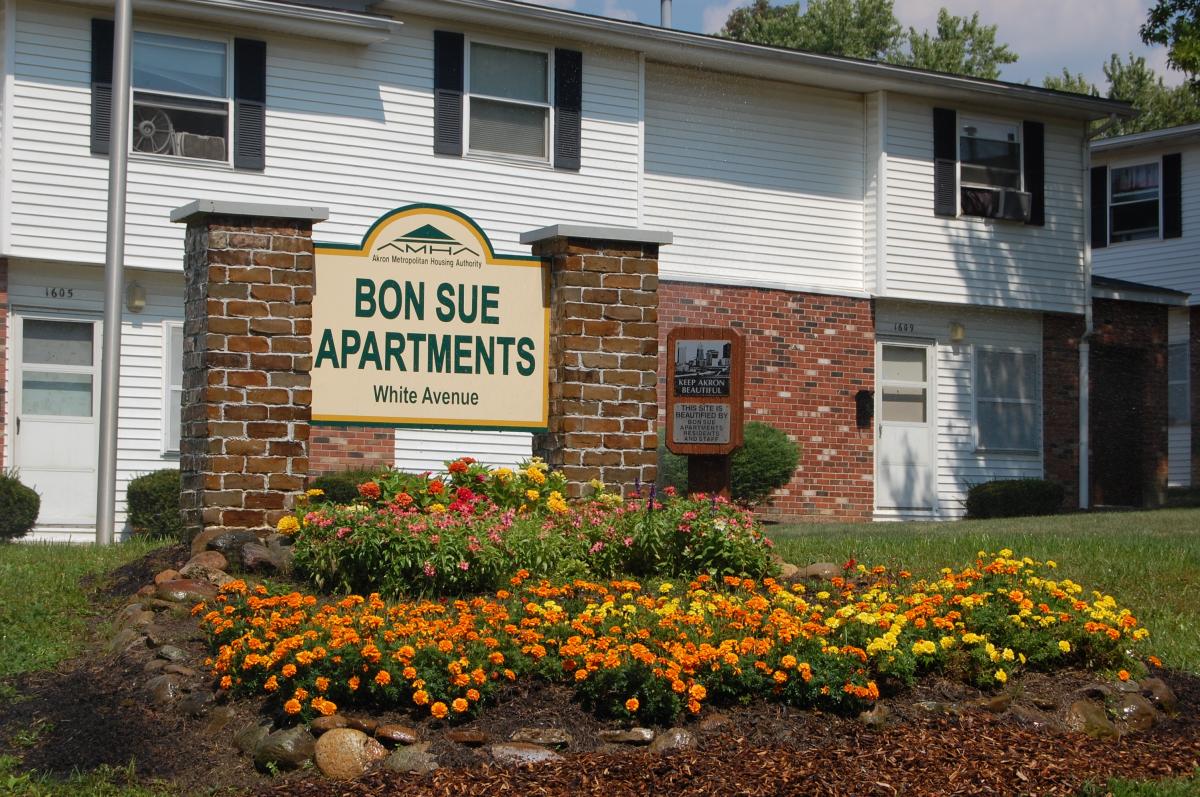 A sign reading "Bon Sue Apartments, White Avenue" stands in front of a brick apartment building with a flower bed and trimmed bushes.