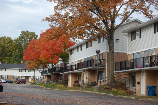Row of townhouse apartments with balconies, bordered by a street and trees with autumn foliage in shades of red and orange.