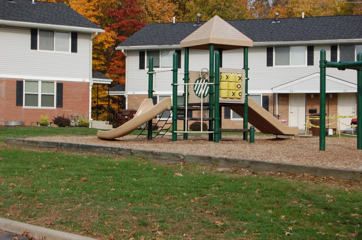 Playground with two slides, climbing bars, and tic-tac-toe panel, situated in front of two-story apartment buildings with autumn trees in the background.