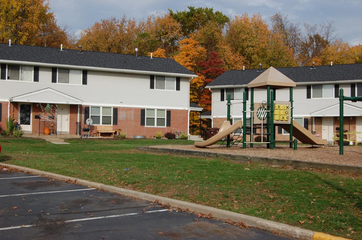 Two-story brick and white siding apartment buildings with a playground featuring slides and tic-tac-toe. Trees with autumn foliage are in the background.