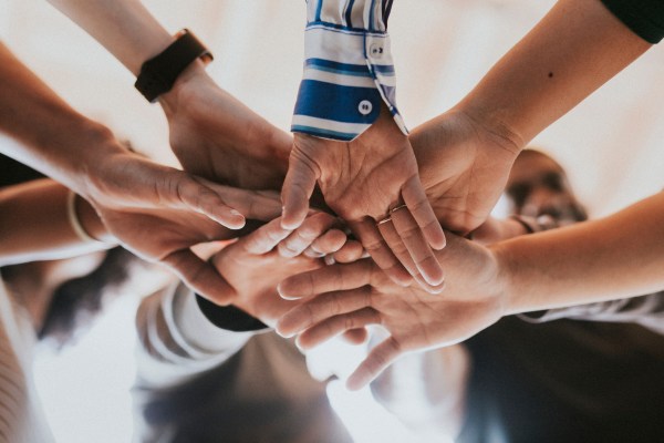 A group of people stack their hands together in the center, viewed from below, symbolizing teamwork and unity.