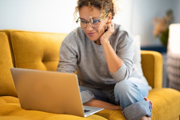A woman wearing glasses sits on a yellow sofa, looking at a laptop on her lap in a well-lit room.