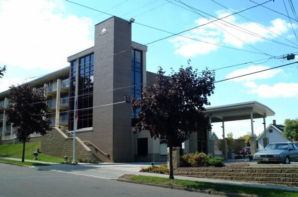 A modern, three-story brick building with large windows and a covered entrance, surrounded by trees, cars, and a sidewalk on a sunny day.
