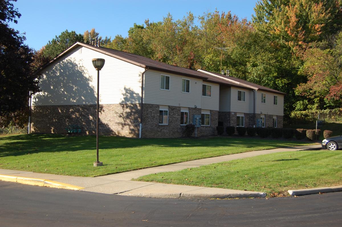 Two-story apartment building with beige siding and brick facade, surrounded by a green lawn, trees, and a sidewalk with a streetlamp.