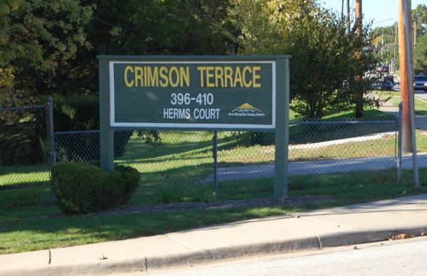 A green sign reads "CRIMSON TERRACE 396–410 HERMS COURT" with a yellow AMHA logo underneath, situated beside a sidewalk and road.