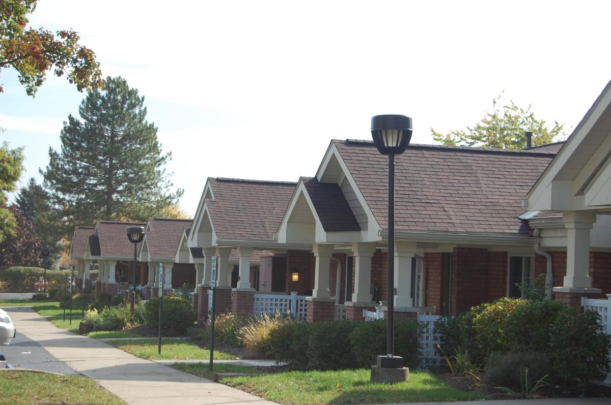 Row of single-story brick houses with front porches, neatly trimmed bushes, and streetlights lining a sidewalk on a sunny day.