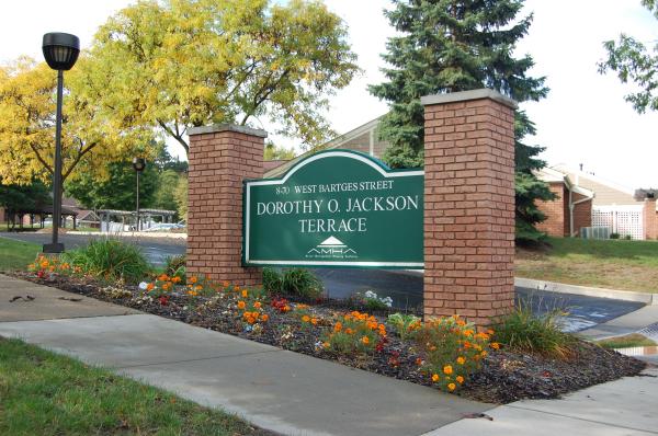 A green sign between brick columns reads "Dorothy O. Jackson Terrace" on West Battges Street, with flowers and trees visible in the background.