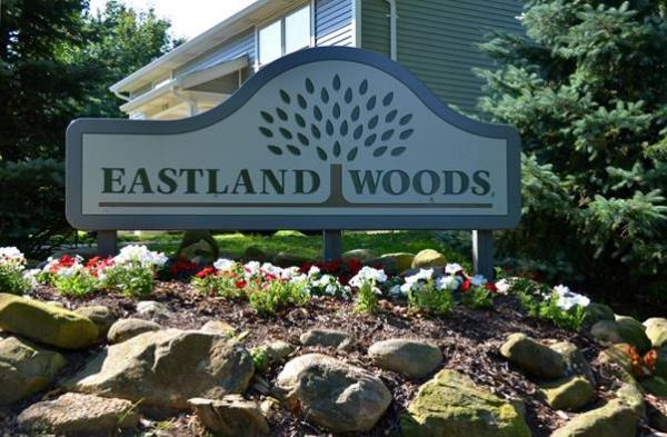 A sign reading "Eastland Woods" with a tree graphic is surrounded by rocks, flowers, and greenery in front of a building.
