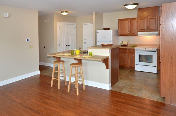 Modern kitchen with wood cabinets, white appliances, breakfast bar with two stools, and hardwood floors adjacent to a tiled area.