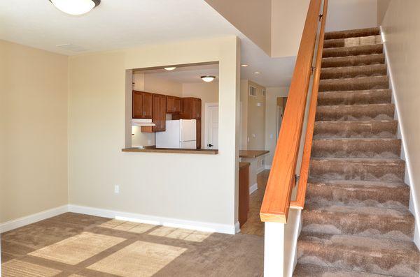 Carpeted stairs and landing, beige walls, and a view into a kitchen with wooden cabinets and white appliances. Natural light coming from a window out of frame.