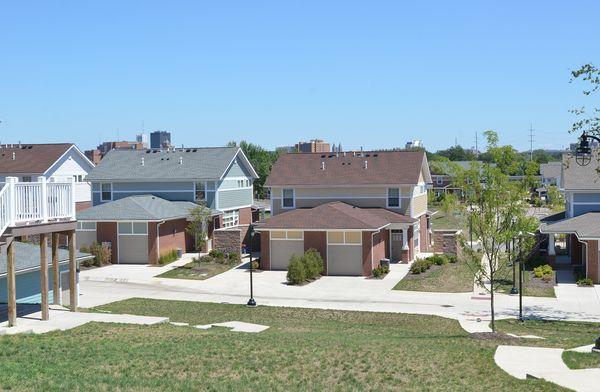 Suburban neighborhood with two-story houses, garages, green lawns, and a clear blue sky in the background.