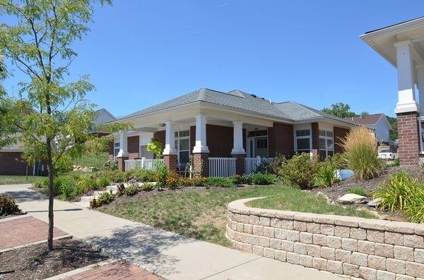 Single-story brick house with a covered front porch, surrounded by landscaped yard, shrubs, a small tree, and a curved stone retaining wall under a clear blue sky.