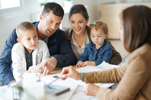 A family with two young children sits at a desk, attentively listening to a professional woman discussing documents in an office setting.