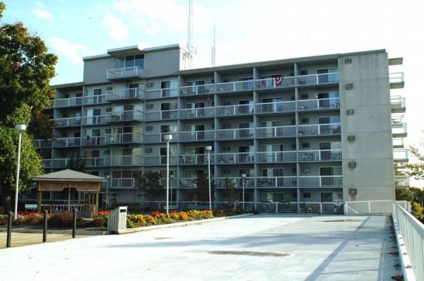 A five-story apartment building with multiple balconies and a parking area in front, adjacent to a small gazebo and landscaped garden area.