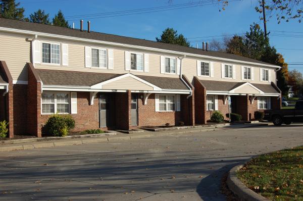 Row of two-story brick and beige townhouses with small bushes in front, concrete driveways, and parked black pickup truck on the right. Trees and power lines visible in the background.