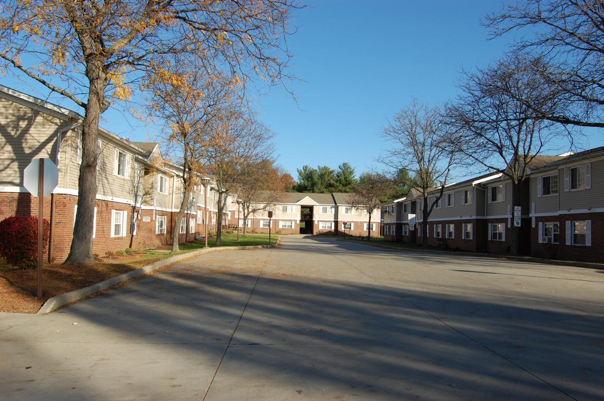 An empty paved street runs between two rows of multi-unit apartment buildings, with bare trees and clear blue sky overhead.