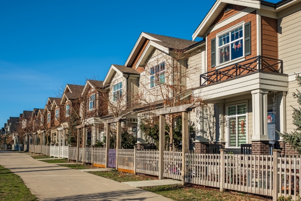 Row of modern two-story houses with fenced front yards along a sidewalk on a clear, sunny day.