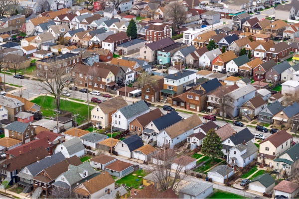 Aerial view of a suburban neighborhood with rows of houses, green lawns, trees, and parked cars along the streets.