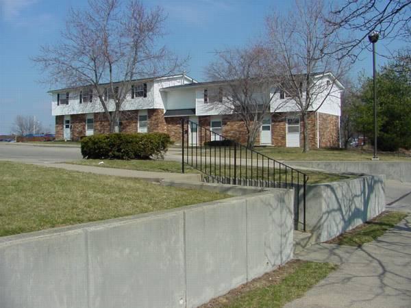 Two two-story apartment buildings with white siding and brick lower walls, separated by a driveway. Leafless trees and a sidewalk are in the foreground.