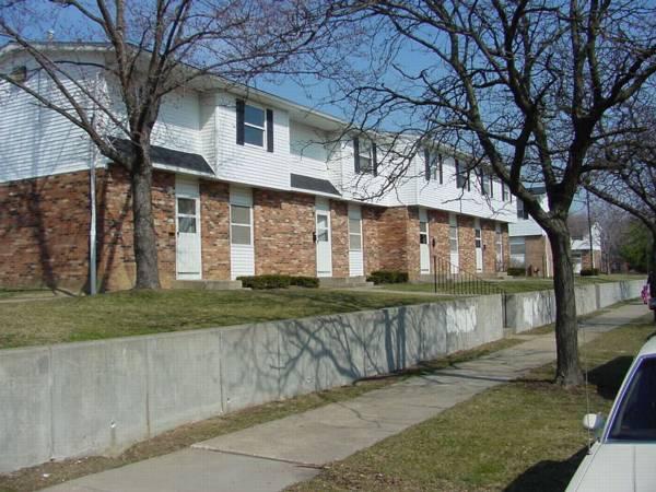 Two-story brick and white townhouse apartments with bare trees, a concrete retaining wall, and a sidewalk in front on a clear day.