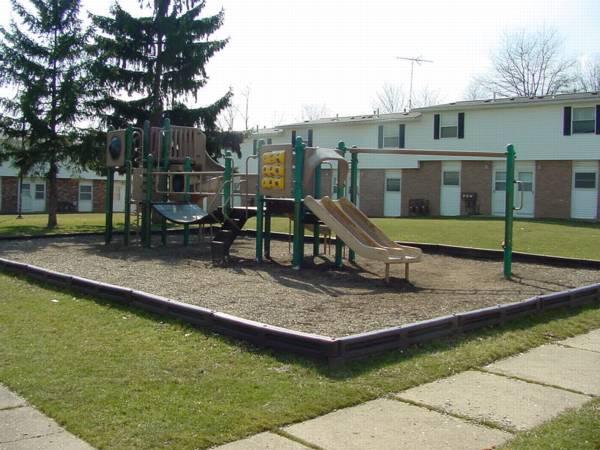 Outdoor playground with slides, climbing structures, and swings, bordered by wooden beams, located in a grassy area near apartment buildings.
