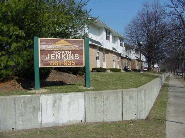 A brown sign reading "North Jenkins 861-935" stands in front of a row of beige and white apartment buildings beside a sidewalk.
