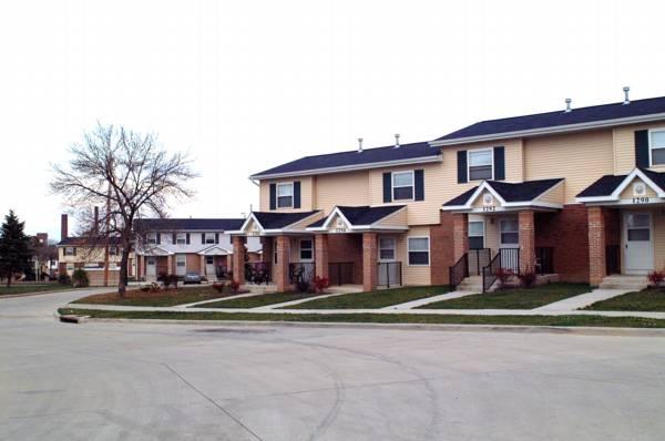 Row of two-story townhouses with beige siding and brick facades, situated along a paved residential street with grass lawns and a few trees.